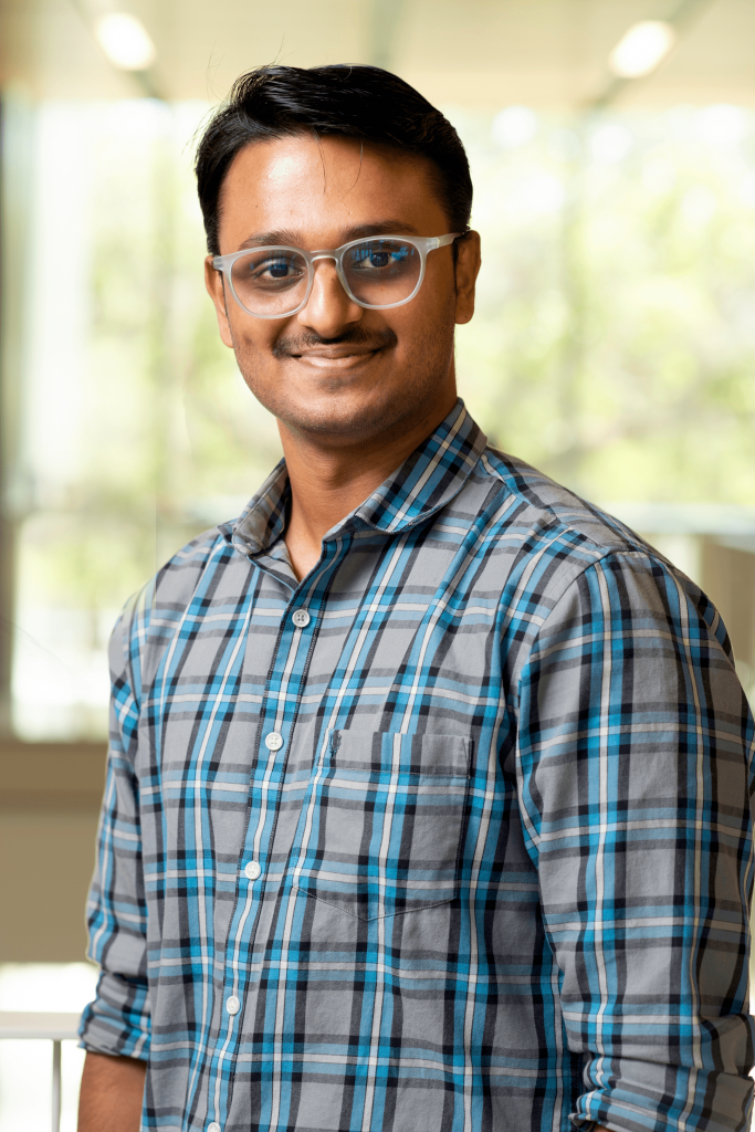 Aniket Malpure, wearing a blue and gray plaid shirt and a wristwatch, smiles while standing on an indoor balcony of the Malachowksy building at UF, with students visible in the background.