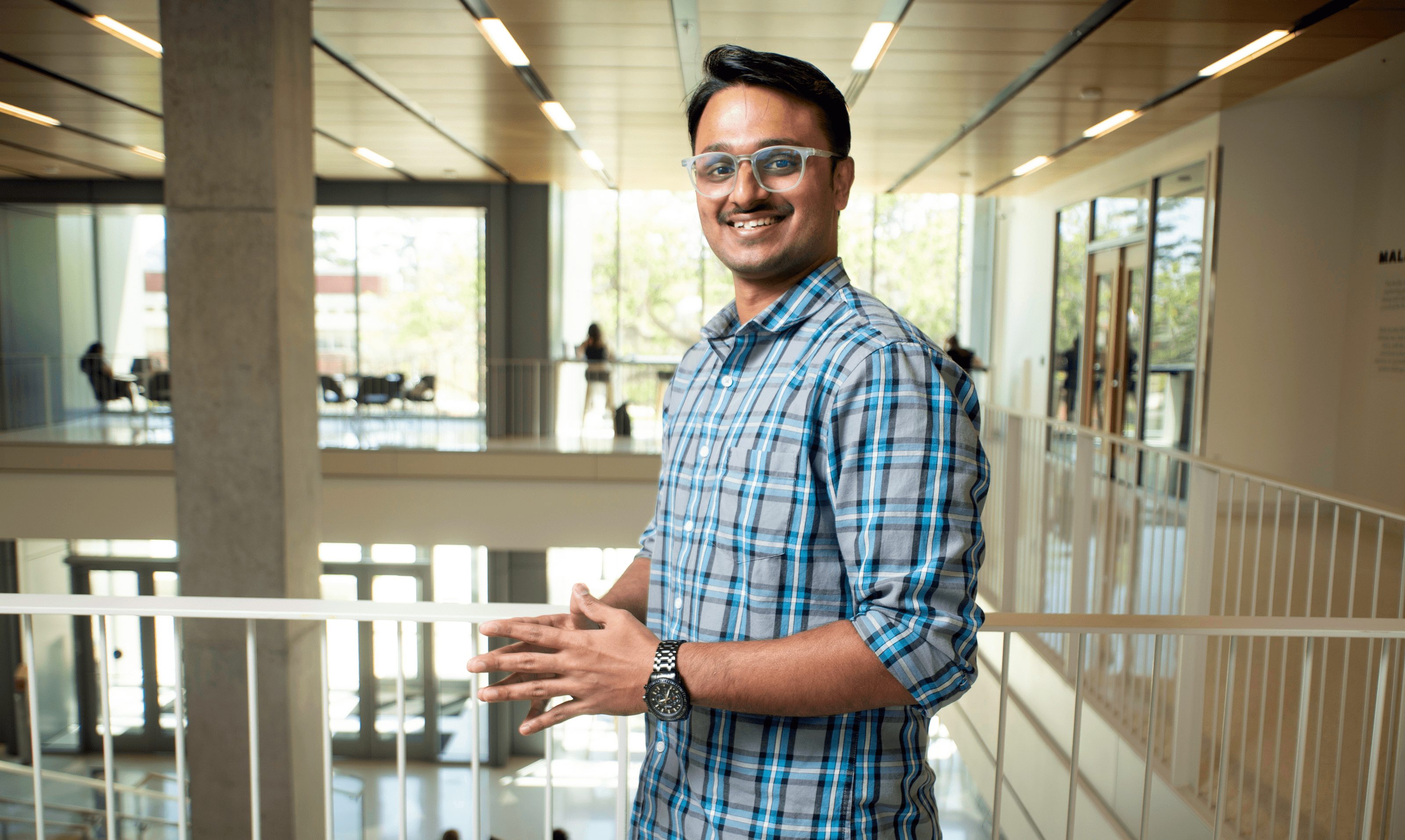 Aniket Malpure, a graduate student with short black hair and glasses, smiles while wearing a blue and gray plaid shirt. He is standing indoors with a softly blurred background of windows and trees.