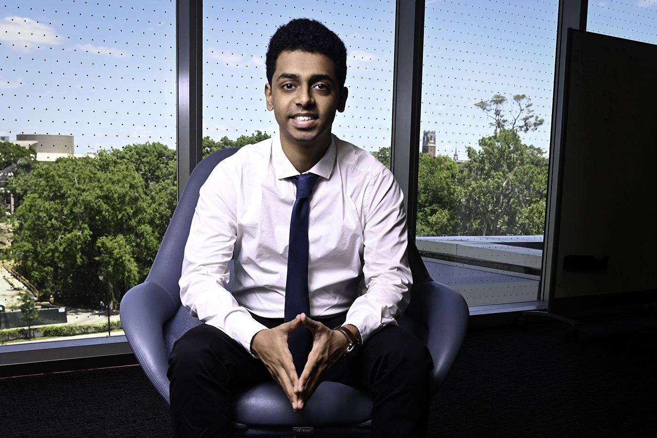 Anshul Patil, a young man in a white shirt and blue tie, seated in a modern chair by a large window overlooking the University of Florida campus. He is smiling and facing the camera with his hands clasped in front of him.